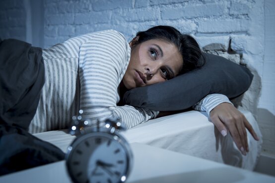 Junge Frau liegt nachts wach im Bett, stützt Kopf auf Kissen, Wecker auf Nachttisch, Symbol für Schlaflosigkeit
