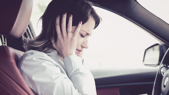 Junge Frau hält sich gestresst den Kopf und schließt die Augen auf dem Fahrersitz eines Autos