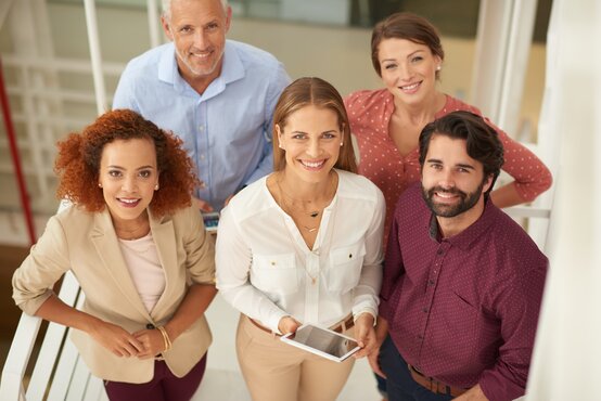 Fünfköpfiges diverses Team in Business-Kleidung steht lächelnd auf weißer Treppe im modernen, hellen Büro