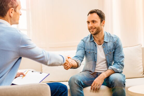 Psychologe oder Arzt im Gespräch mit Patient, beide sitzen entspannt in heller Praxis und reichen sich die Hand