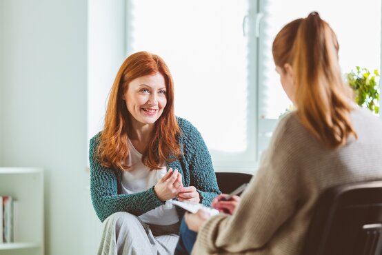 Zwei Frauen sitzen sich in hellem Raum gegenüber, eine lächelt und gestikuliert, die andere schreibt auf Block