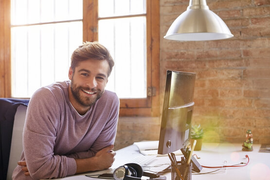 Junger Mann mit Bart sitzt lächelnd am Schreibtisch im modernen, hellen Büro mit Computer und Backsteinwand