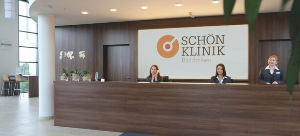 Moderner Empfangsbereich der Schön Klinik Bad Arolsen mit drei Mitarbeiterinnen und leuchtendem Klinik-Logo