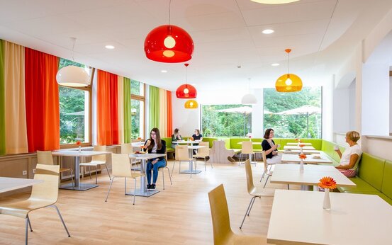 Heller Speisesaal der Klinik mit modernen Holzmöbeln, großen Fenstern und farbenfrohen Vorhängen