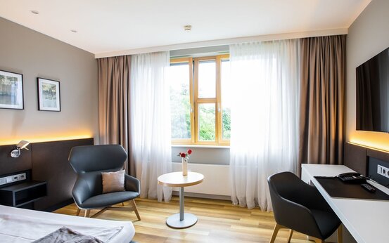 Modernes Einzelzimmer mit großem Fenster, grauem Sessel, Holztisch mit Blumen, Schreibtisch, Flachbildfernseher und heller Einrichtung