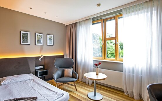 Modernes Einzelzimmer mit Bett, Nachttisch, Sessel, rundem Tisch mit Vase und Rosen, großem Fenster und Holzfußboden