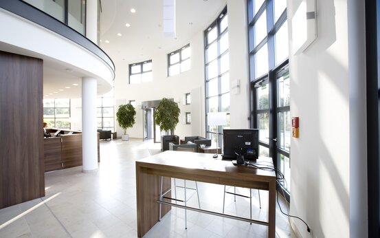 Helles, modernes Foyer mit bodentiefen Fenstern, dunklem Empfangstisch, Sitzgruppe, Zimmerpflanzen und Drehkreuz-Eingang