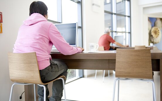 Heller Eingangsbereich mit großen Fenstern, Holzmöbeln und zwei Personen an langen Tischen mit Laptops