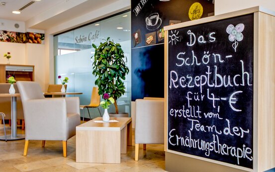 Gemütliches Klinikcafé mit hellen Sitzmöbeln, Blumen, Kreidetafel zum Schön-Rezeptbuch und großer Zimmerpflanze im Tageslicht