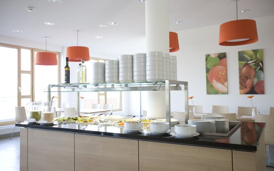 Modernes, heller Speisesaal mit Buffet, frischen Salaten, Getränken und Sitzbereich mit Holzmöbeln und großen Fenstern