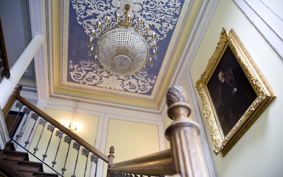 Elegantes historisches Treppenhaus mit Holzhandlauf, verziertem Metallgeländer, Kristallkronleuchter und goldgerahmtem Porträtgemälde