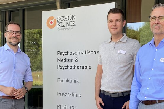 Drei Männer mit Namensschildern vor dem Eingang der Schön Klinik Bad Bramstedt, Kliniklogo sichtbar