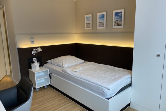 Modernes Einzelzimmer in Klinik mit Einzelbett, Nachttisch, Kleiderschrank und drei Landschaftsbildern an der Wand