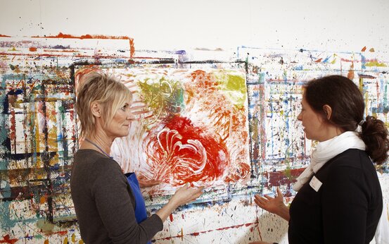 Zwei Frauen im Atelier stehen vor abstrakten, farbenfrohen Gemälden während der Kunsttherapie