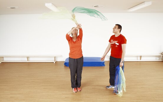 Ältere Frau wirft bunte Gymnastiktücher bei Bewegungstherapie, Mann lächelt, blaue Matten im Hintergrund