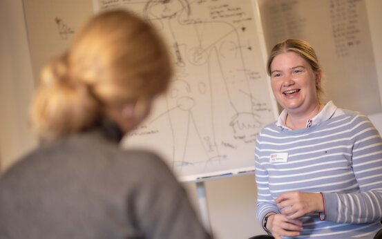 Blonde Therapeutin spricht freundlich mit Patientin vor Flipchart mit Anatomiezeichnung in medizinischer Praxis