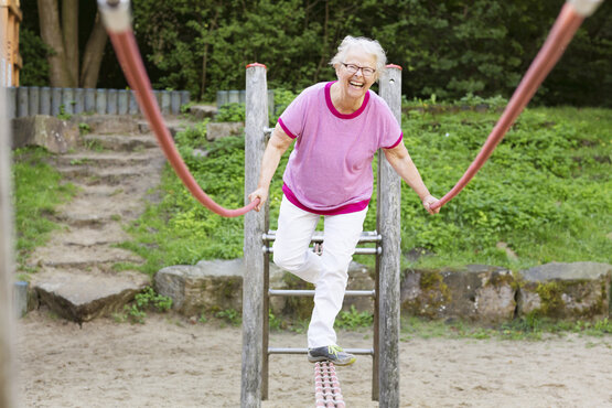 Ältere Frau balanciert lächelnd auf Trainingsgerät im Freien, hält sich an roten Seilen fest