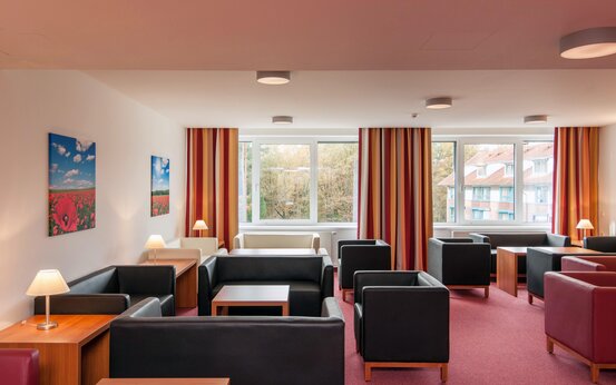 Heller Aufenthaltsraum mit schwarzen und weißen Ledersesseln, Holztischen, großen Fenstern und rot-gelb gestreiften Vorhängen