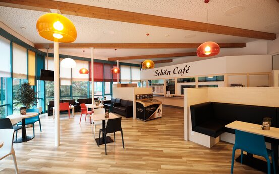 Modernes, helles Café „Schön Café“ mit bunten Stühlen, Holztischen und großen Fenstern