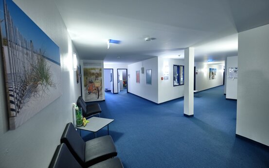 Flur in Klinik mit Sitzgruppe, blauem Teppich, Naturfotos und offenen Türen zu Behandlungsräumen