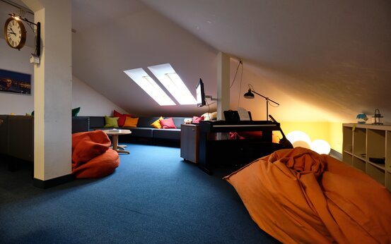 Gemütlicher Aufenthaltsraum im Dachgeschoss mit Sitzecke, bunten Kissen, Digitalpiano und Tageslicht durch Dachfenster