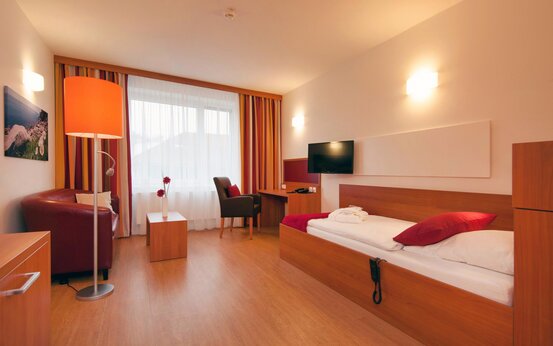 Modernes, barrierefreies Patientenzimmer mit Einzelbett, Sessel, Schreibtisch, großem Fenster und Holzfußboden in Klinik