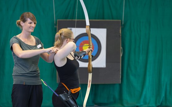 Zwei Frauen beim Bogenschießen-Training in Halle, Trainerin korrigiert Schusshaltung vor bunter Zielscheibe