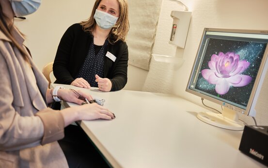 Patientin mit Sensoren an Fingern bei Biofeedback-Therapie, Therapeutin am Tisch, Monitor zeigt rosa Lotusblüte