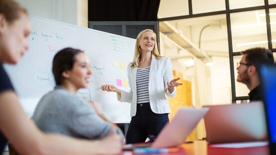 Vier Personen bei Teammeeting im modernen Büro, Frau präsentiert am Whiteboard, Fokus auf Zusammenarbeit