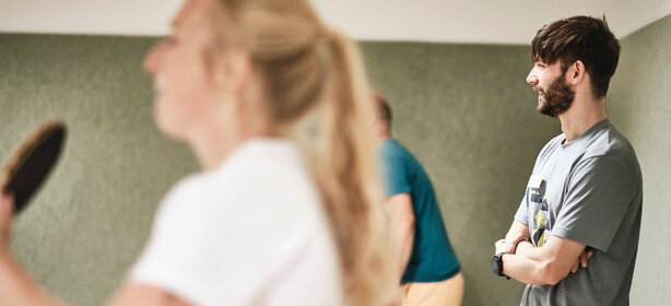 Junger Mann mit Bart beobachtet lächelnd ein Tischtennisspiel im Indoor-Freizeitbereich 