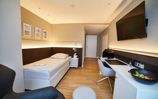 Modernes, helles Einzelzimmer mit Einzelbett, Schreibtisch, Flachbildfernseher und Ledersessel