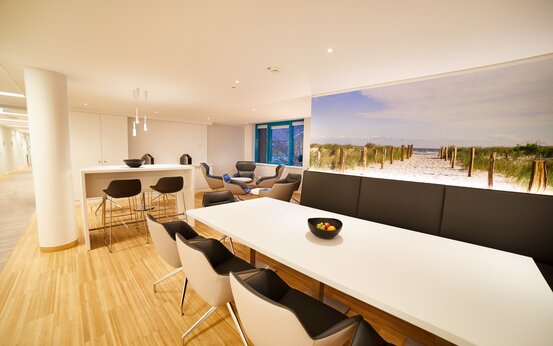 Heller Aufenthaltsraum mit großem weißen Tisch, Designerstühlen, Obstschale, Küchenzeile und großem Dünenbild an der Wand