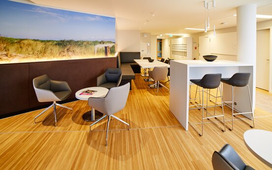 Heller Aufenthaltsraum in Klinik mit gepolsterten Sesseln, runden Tischen, Theke und großem Dünenfoto an der Wand