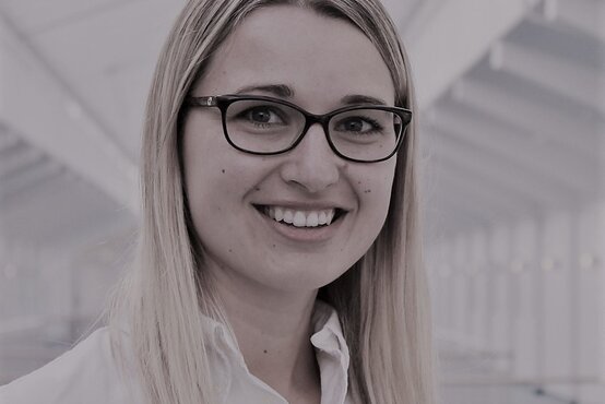 Tessa Schneeberger. Junge Frau mit langen Haaren und Brille lächelt in moderner, heller Geschäftsumgebung in die Kamera