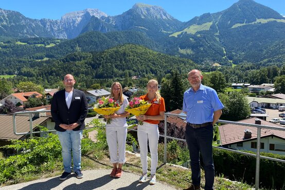 Zwei ausgezeichnete Auszubildende mit Blumensträußen und zwei Gratulanten vor Alpenkulisse, Ehrung im Gesundheitswesen