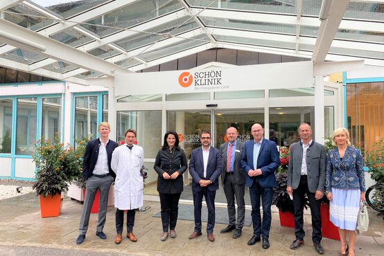 Acht Personen, darunter Arzt im weißen Kittel, Klinikleitung und Gäste, vor Haupteingang der Schön Klinik Berchtesgadener Land bei Tageslicht