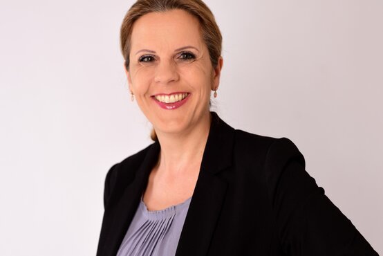 Astrid Angele. Porträt einer lächelnden Frau mit blonden, zurückgebundenen Haaren, schwarzem Blazer und hellgrauer Bluse