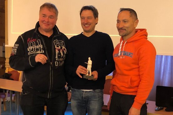 Drei Männer in Klinik-Seminarraum, mittlerer Mann hält Auszeichnung