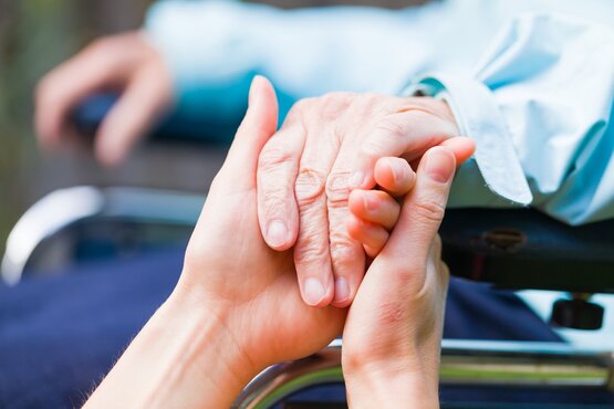 Nahaufnahme: Jüngere Hand hält unterstützend die Hand einer älteren Person im Rollstuhl