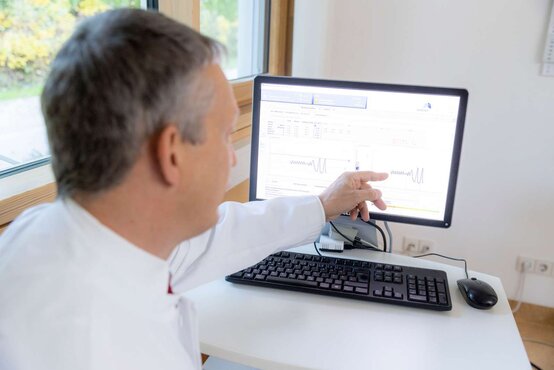 Arzt im weißen Kittel zeigt in heller Praxis auf Computerbildschirm mit medizinischen Diagnosedaten und Kurvendiagrammen