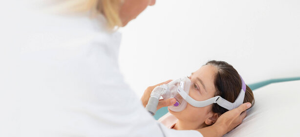 Ärztin legt Patientin auf Klinikbett fürsorglich eine Atemmaske an, helle Umgebung