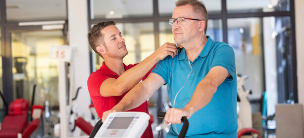 Mann mit Sauerstoff-Nasenbrille trainiert auf Ergometer, Physiotherapeut überwacht Vitalwerte