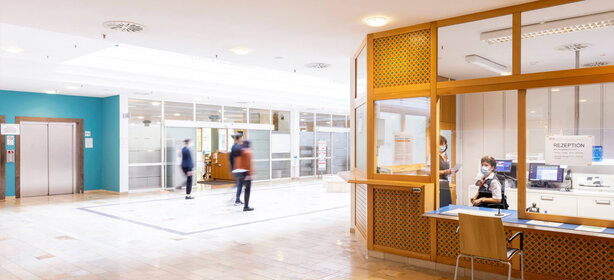 Heller Klinik-Eingangsbereich mit Empfangstresen, zwei Mitarbeitenden mit Mundschutz, Aufzug und klarer Beschilderung
