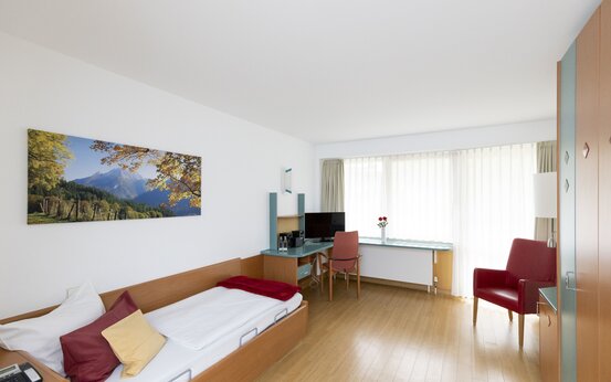 Helles Einzelzimmer mit Pflegebett, Schreibtisch, Sessel, großem Fenster und moderner Ausstattung