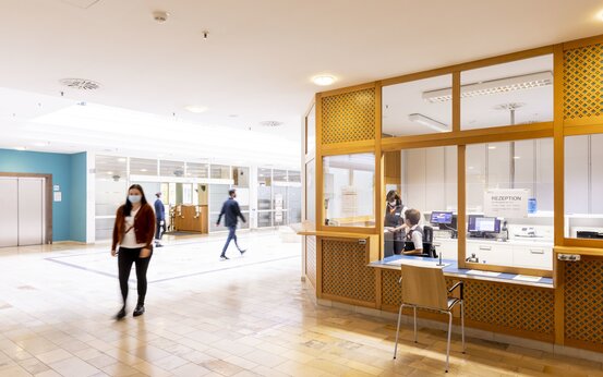 Helle Krankenhaus-Lobby mit moderner Rezeption, zwei Mitarbeiterinnen am Computer, Personen mit Mundschutz im Empfangsbereich