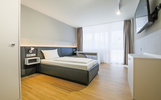Modernes Einzelzimmer mit großem Fenster, Einzelbett, Schreibtisch, TV und heller, komfortabler Einrichtung