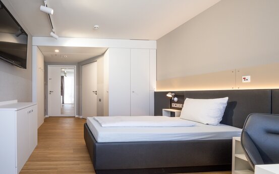 Einzelzimmer mit Holzboden, großem Bett, Schreibtisch, TV und heller, komfortabler Atmosphäre