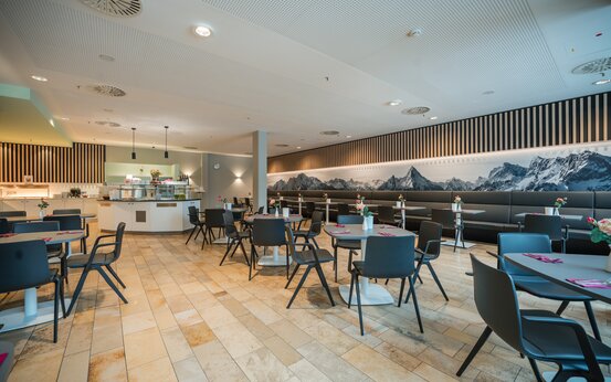 Moderner, heller Speisesaal mit stilvollen Sitzgruppen, Blumenvasen und großem Bergpanorama an der Wand