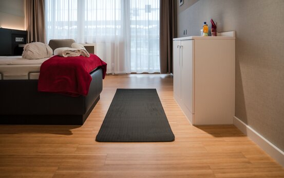 Modernes, helles Zimmer mit Bett, roter Decke, weißen Handtüchern, Sideboard mit Getränken und großem Fenster