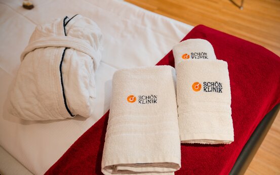 Weißer Bademantel und drei gerollte Handtücher mit Kliniklogo auf roter Decke im modernen Patientenzimmer